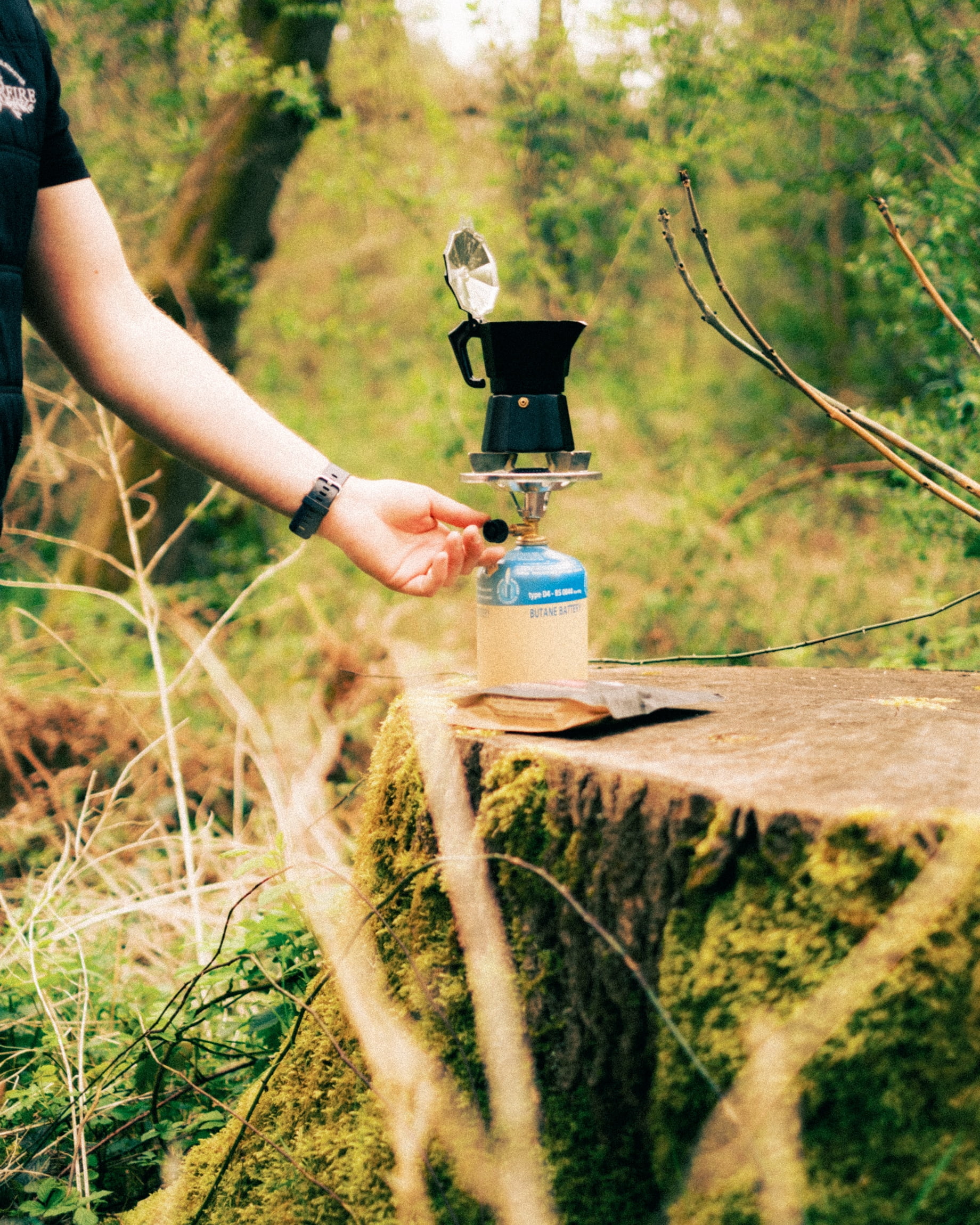 A moka pot sat on a gas stove, which is being lit, on a tree trunk
